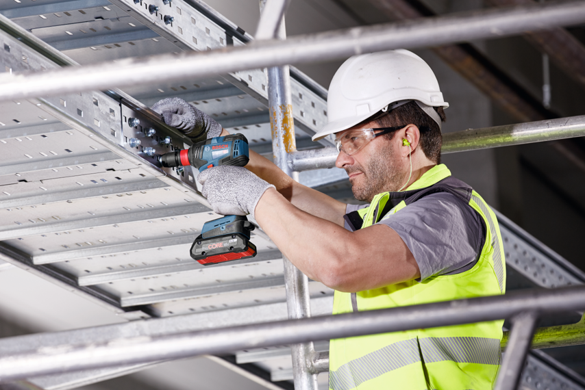 A man in a helmet and gloves uses a Bosch drill on metal scaffolding.