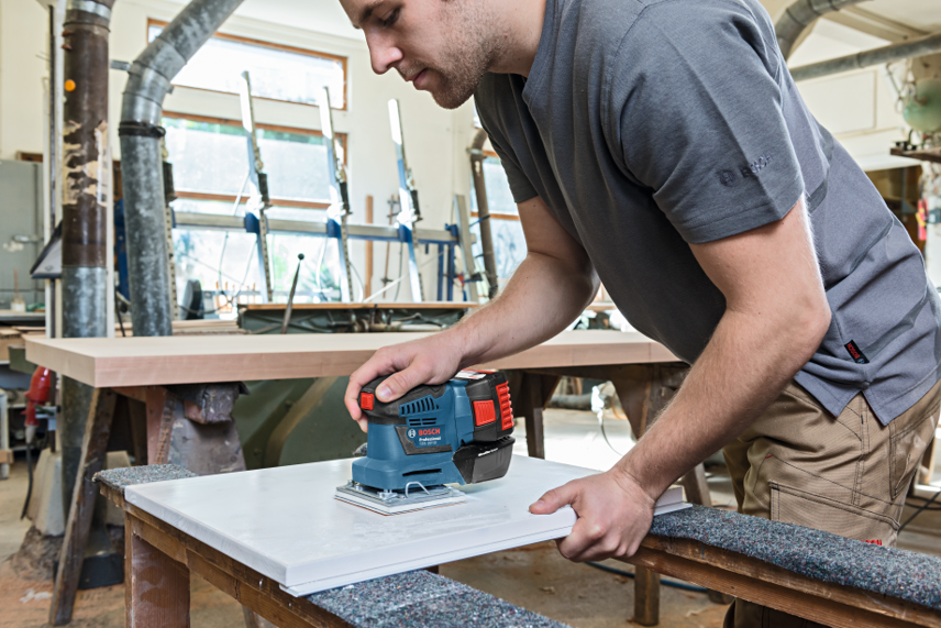 A man uses a Bosch Professional sander on a white board in a workshop.
