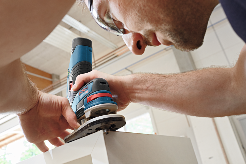 A man uses a blue Bosch Professional router on a white workpiece in a workshop.