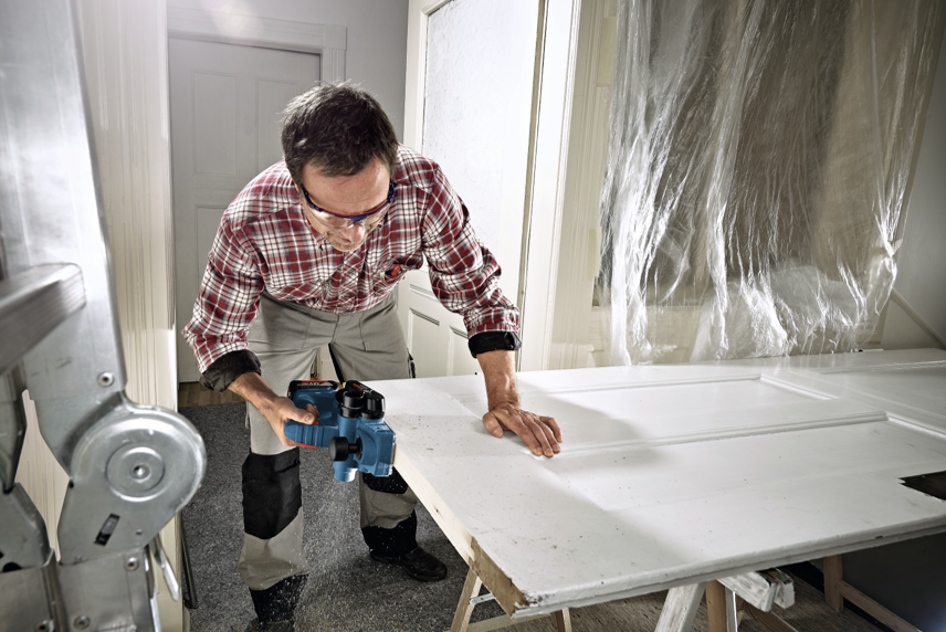 A man in a checked shirt uses a blue Bosch tool on a white door in a room.