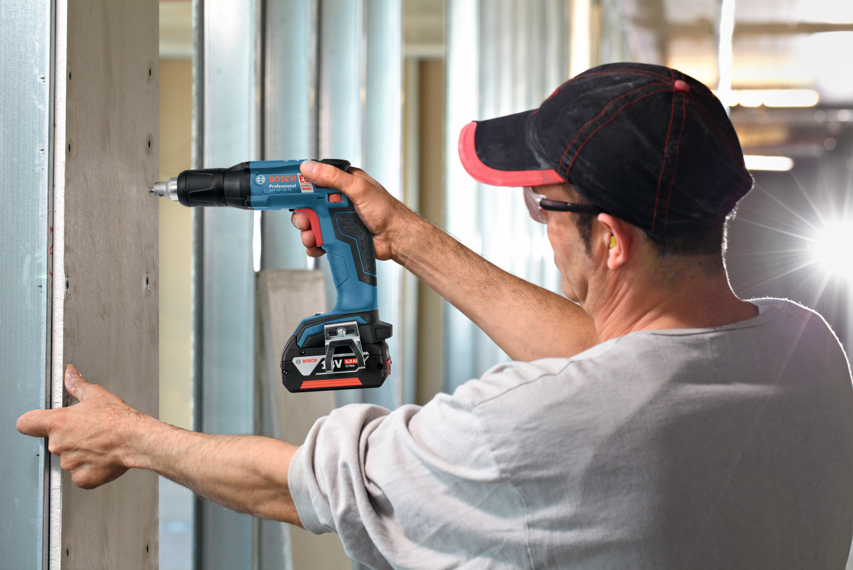 A man with glasses uses a blue Bosch Professional screwdriver on a wall.