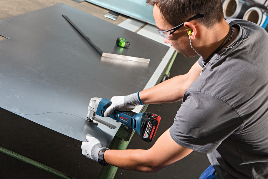 A man uses a blue Bosch Professional jigsaw to cut metal on a workbench.