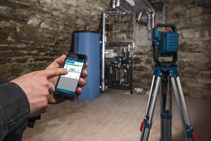 A person uses a smartphone near a blue tank and a Bosch Professional laser level.