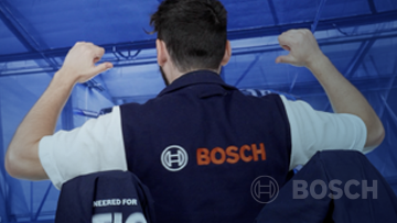 A man in a dark blue vest with the Bosch logo raises his arms in a blue industrial environment.