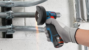 A Bosch Professional cordless angle grinder is cutting through metal pipes on a wall, with sparks flying.