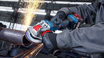 A Bosch Professional cordless angle grinder is cutting through a metal pipe, creating sparks. The tool is blue and red, held by a gloved hand in a workshop setting.