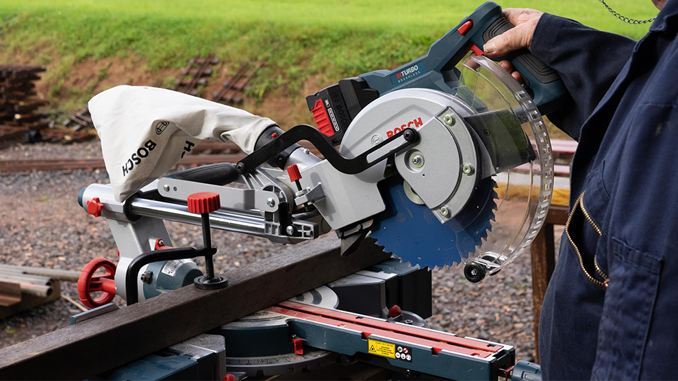 A Bosch Professional miter saw held by a person, cutting a wooden beam outdoors.