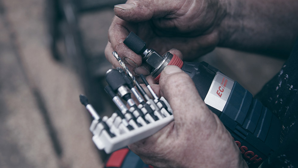 A person holds Bosch tools, including a drill and various bits, in a workshop setting.
