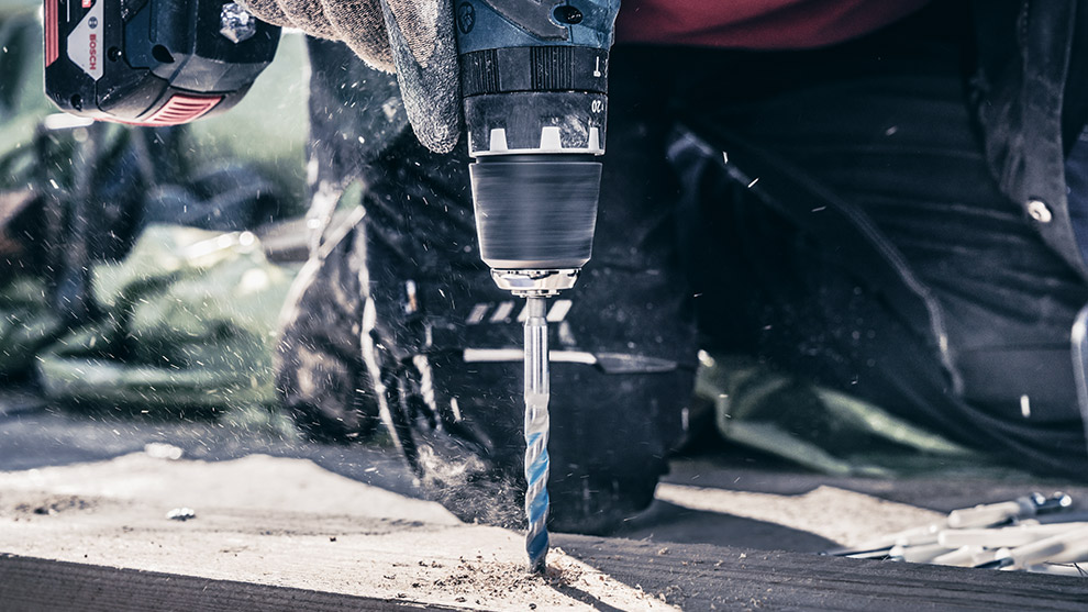 A Bosch Professional drill is powering through wood, with dust flying and a work glove visible.