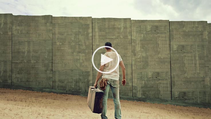 A man in a white shirt and jeans stands with a suitcase before a concrete wall.