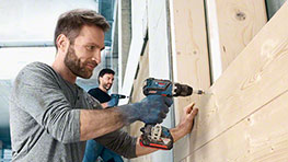 A man using a Bosch Professional drill on wooden boards, another man in the background.