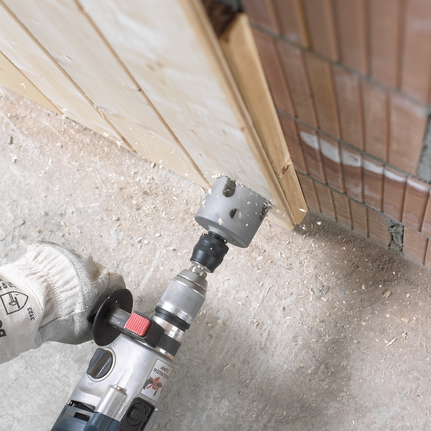 Worker wearing safety equipment drills a hole with a hole saw in a wooden board.