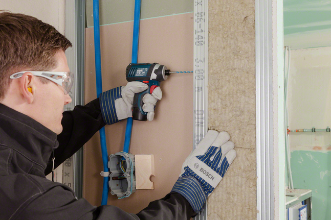 A worker wearing safety equipment drills into drywall beside exposed insulation.