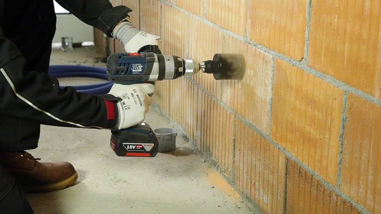 Worker wearing safety equipment drills a hole into a brick wall with a cordless tool.