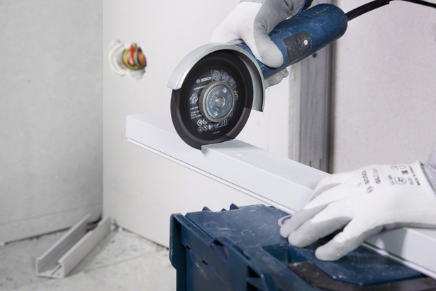A worker wearing safety equipment cuts a metal channel with an angle grinder.