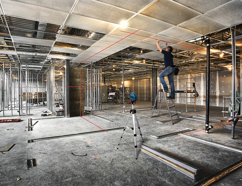 A person on a ladder adjusts ceiling panels in a large room using a laser leveling tool.