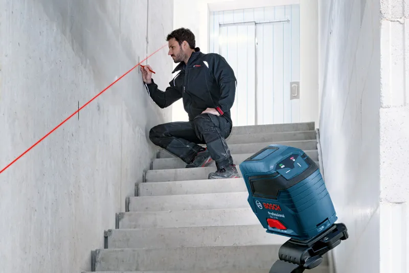 A person wearing safety equipment marks a wall using a laser leveling tool on stairs.