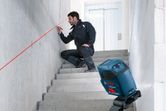 A person wearing safety equipment marks a wall using a laser leveling tool on stairs.