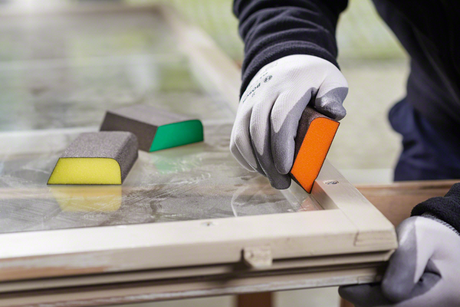 Worker wearing safety equipment sanding a window frame with a sanding sponge.