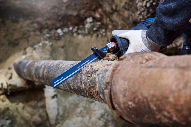 Person wearing safety equipment cuts a rusted metal pipe with a reciprocating saw.