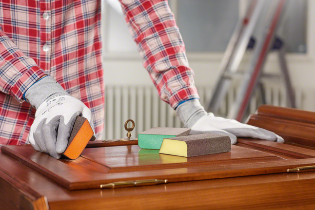 Person wearing safety equipment sanding a wooden cabinet with sanding sponges.