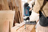 A person wearing safety equipment drills into a wooden plank in a workshop.