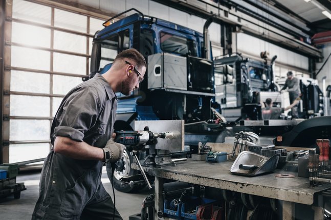 A worker wearing safety equipment drills a hole into metal at a truck workshop.