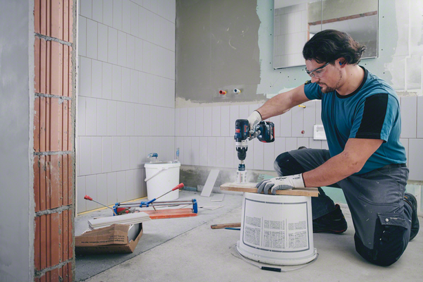A worker wearing safety equipment drills a hole through wood in a partially tiled room.