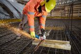 A worker wearing safety equipment cuts rebar on a construction site with an angle grinder.
