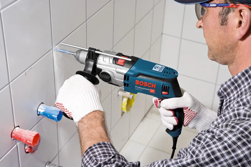 A person wearing safety equipment drills into a tiled wall with an impact drill.