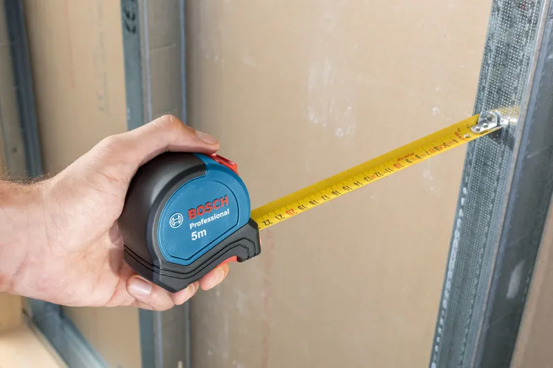 A person uses a tape measure to check distance between drywall and metal frame.