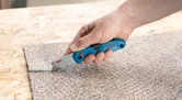 A person uses a folding knife to cut through a piece of carpet.