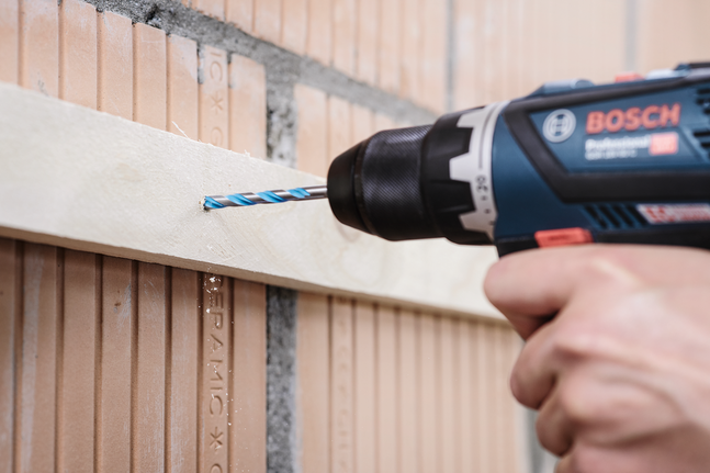 Person drills into a wooden plank mounted on a brick wall.