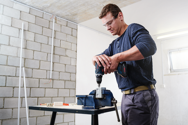 Person wearing safety equipment uses a power drill on metal clamped in a vice.