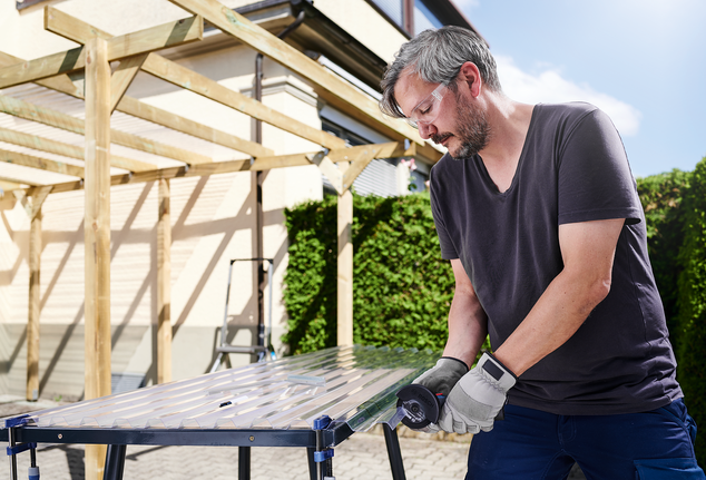 Person wearing safety equipment cuts a clear plastic panel with a power tool outdoors.