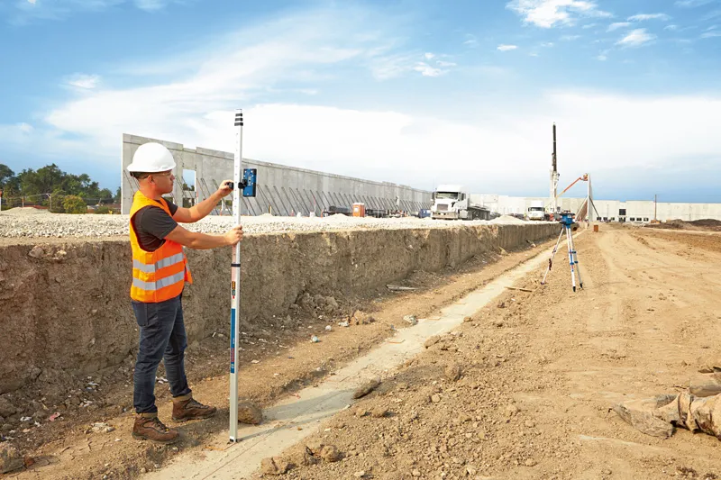 A person wearing safety equipment uses a laser receiver to measure ground level at a construction site.