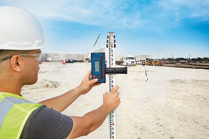 A person wearing safety equipment aligns a laser receiver on a leveling staff at a construction site.