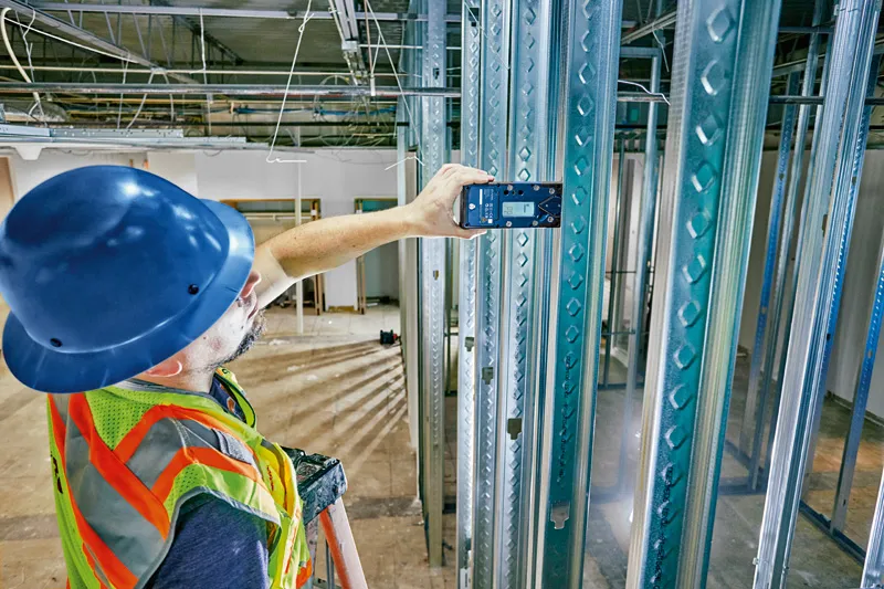 A person wearing safety equipment uses a laser receiver to align metal framing studs.