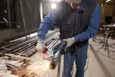 A person wearing safety equipment cuts a metal pipe with an angle grinder, sparks flying.