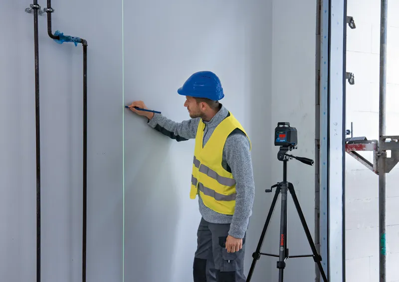 A person wearing safety equipment marks a wall guided by a laser leveling tool on a tripod.