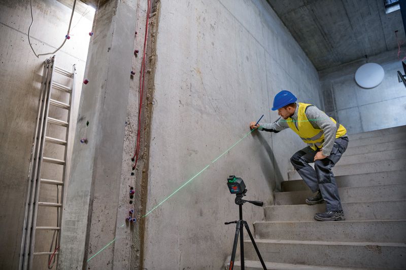 A person wearing safety equipment marks a wall on stairs using a laser leveling tool.