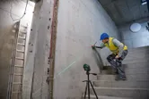 A person wearing safety equipment marks a wall on stairs using a laser leveling tool.