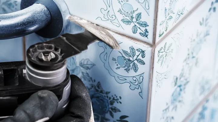 A person wearing safety equipment uses a power tool to remove grout between wall tiles.