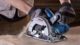 A person wearing safety equipment operates a hand-held circular saw on wood.