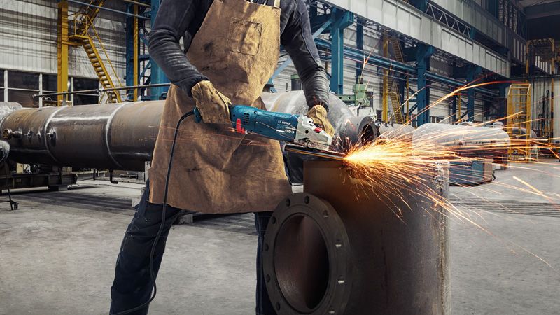 A person wearing safety equipment grinds a metal pipe with an angle grinder, sparks flying.