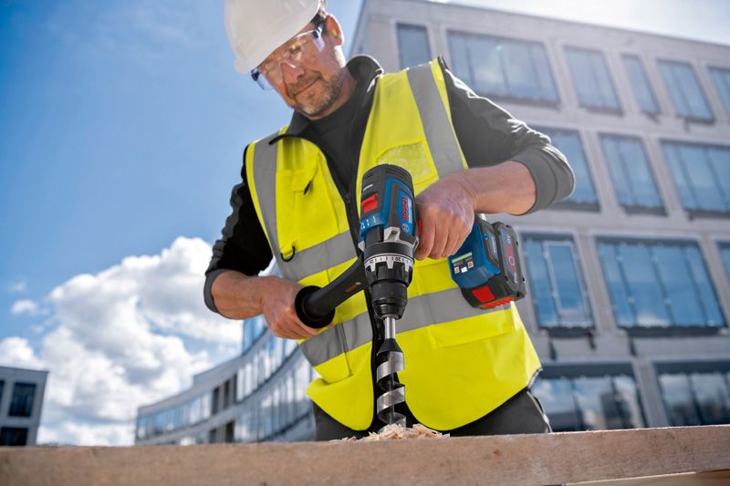 A person wearing safety equipment drills a large hole in wood with a cordless tool.