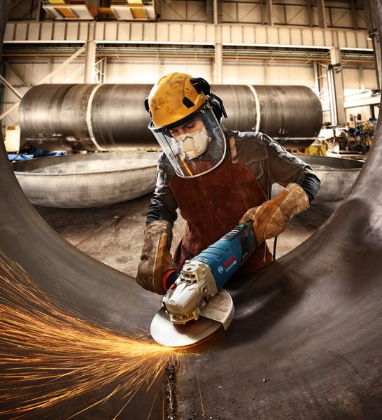 A person wearing safety equipment grinds a large metal pipe with sparks flying.