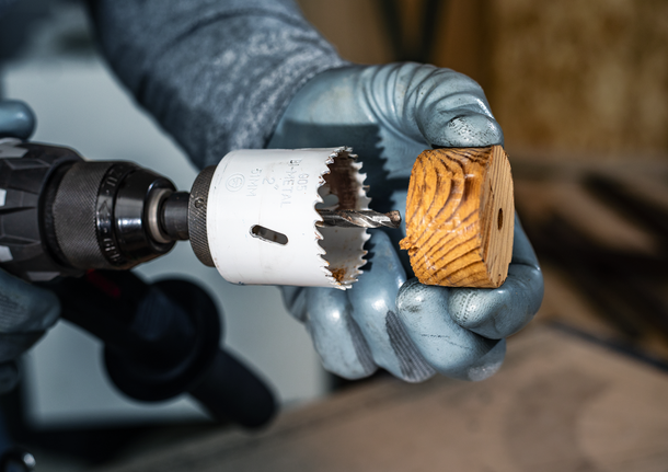 Person wearing safety equipment holds a hole saw drill and a round wooden block.