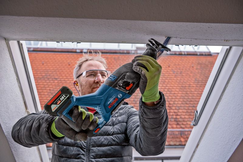 A person wearing safety equipment uses a cordless reciprocating saw to cut a window frame.