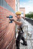 A person wearing safety equipment drills into a brick wall on scaffolding.